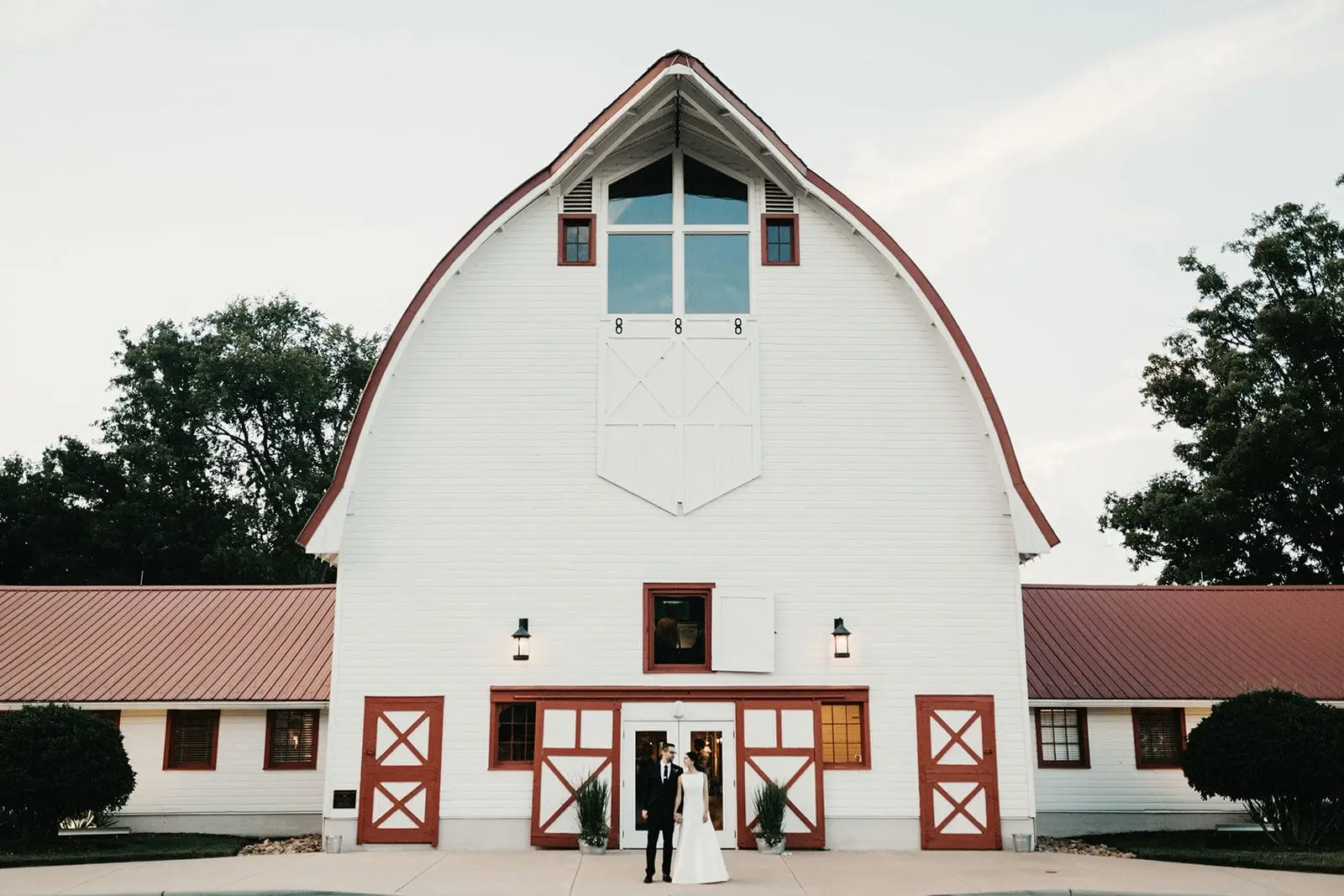 The white Main Barn at WinMock with its red doors at dusk, a couple at the entrance